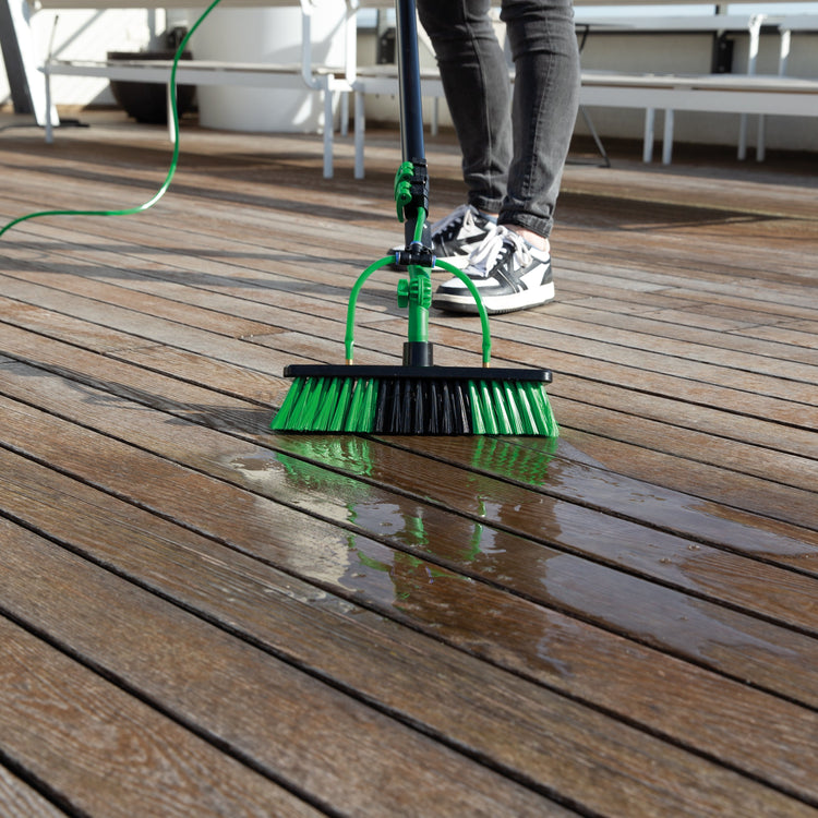 Person cleaning a wooden deck with a green broom and hose.
