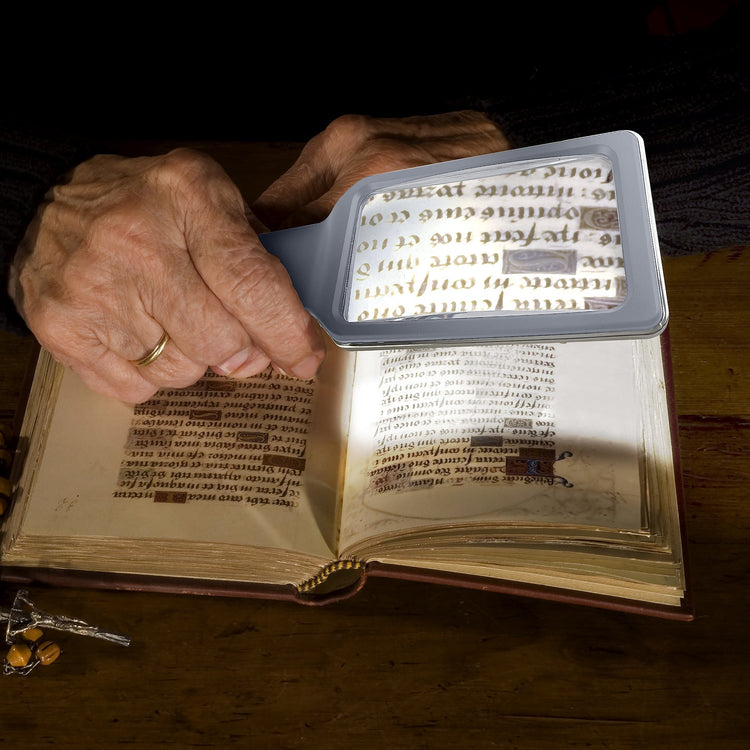 Person using a magnifying glass to read an open book on a wooden surface