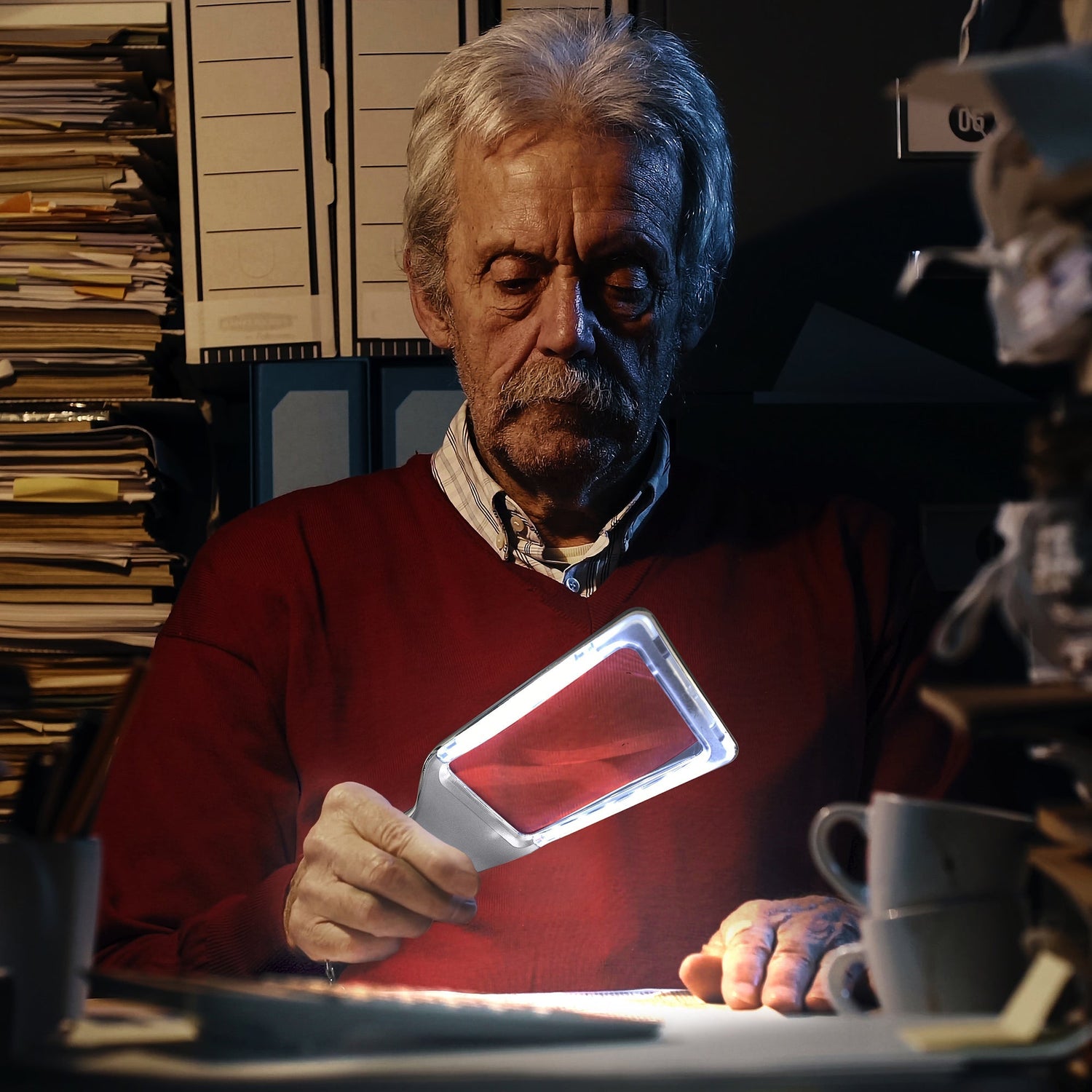 Man using a lamp to read a document in a cluttered office.