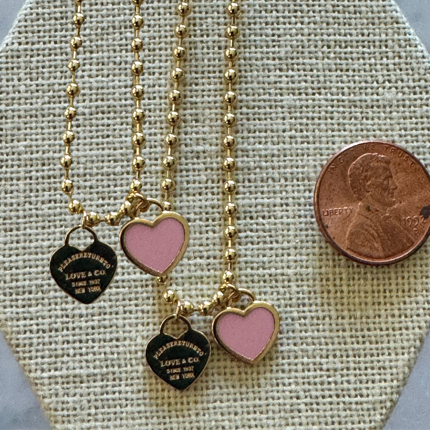 Two gold necklaces with heart-shaped charms on a textured surface, one pink and one black, with a penny for scale.