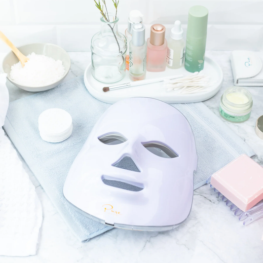 A LED light therapy facial mask and a bottle of high energy hyaluronic serum, both branded by Pure Daily Care, with the mask illustrated in a rainbow of light colors and the serum bottle labeled.