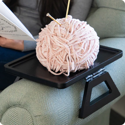Ball of yarn with knitting needles on a small table next to a person reading a book.