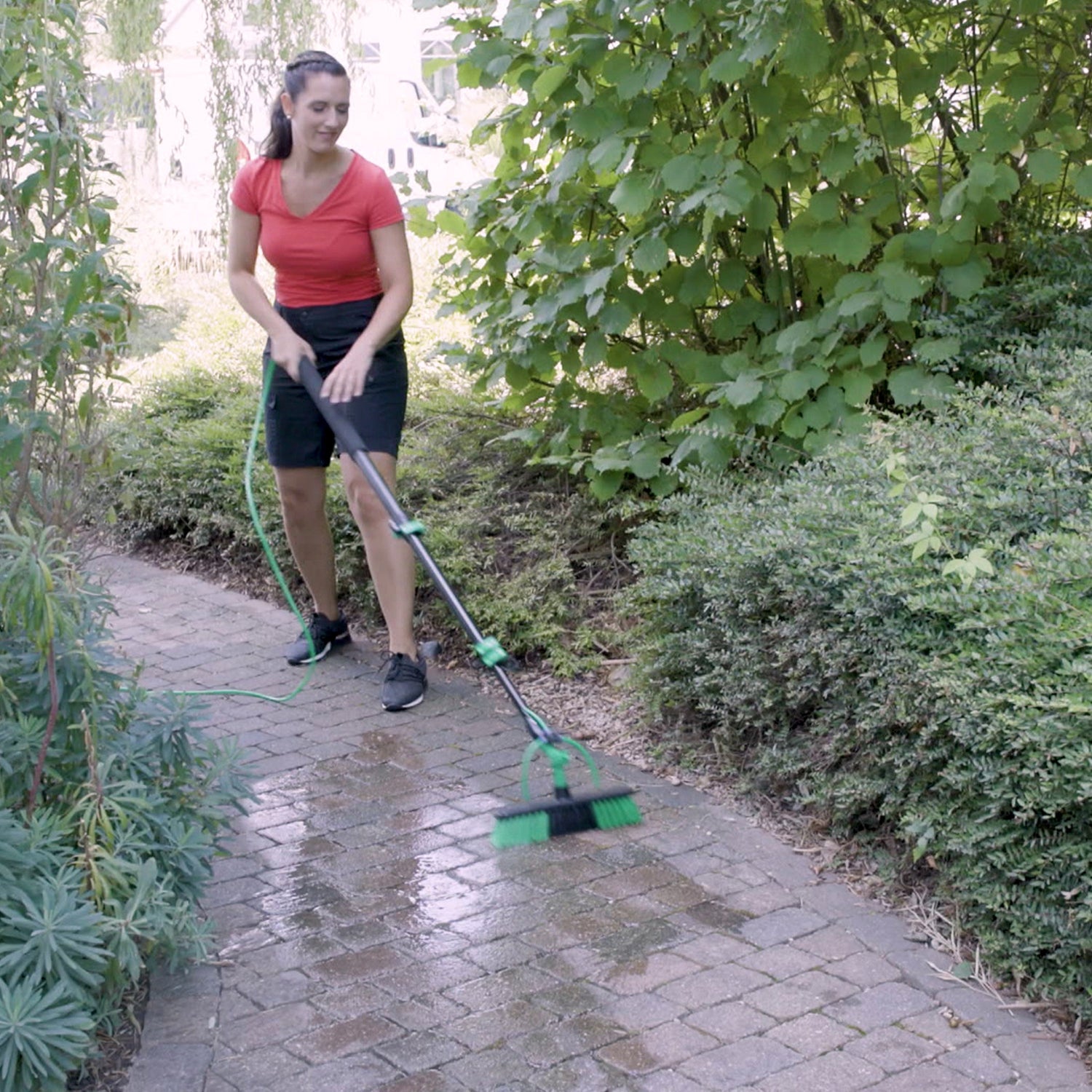Person using a garden broom on a paved path surrounded by greenery